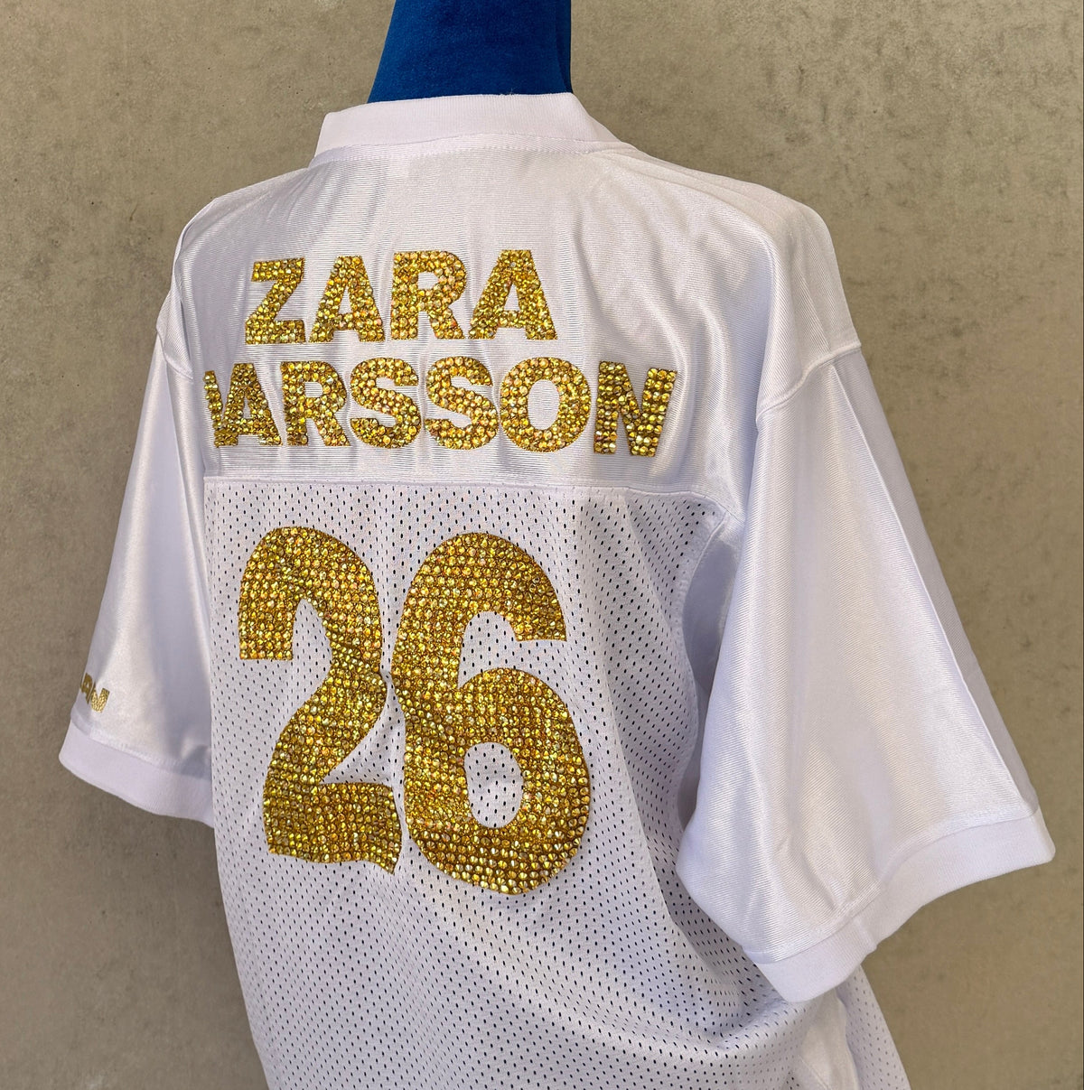 White mesh sports jersey displayed on a mannequin torso, showing the back with 'ZARA LARSSON' and the number '26' in gold lettering, against a textured beige wall.